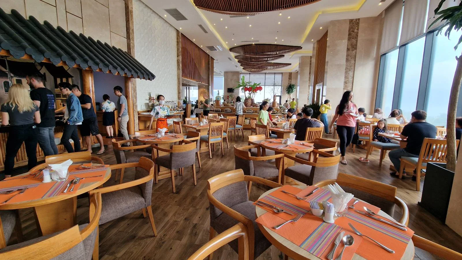 A spacious restaurant with wooden floors and tables, set with orange placemats. Large windows let in natural light. Several people are dining or walking around, and there is a buffet area in the background.