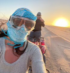 A person wearing goggles and a bandana taking a selfie with another individual on a motorcycle in the background during sunset in a dusty environment.