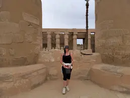 A person stands between two ancient stone columns at the historic Karnak Temple, wearing a black tank top, cap, and sunglasses. Stone ruins and a palm tree are visible in the background under a cloudy sky.