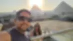Man and woman wearing sunglasses smiling on a rooftop with Giza pyramids in background at sunset. Bright sky and relaxed mood.