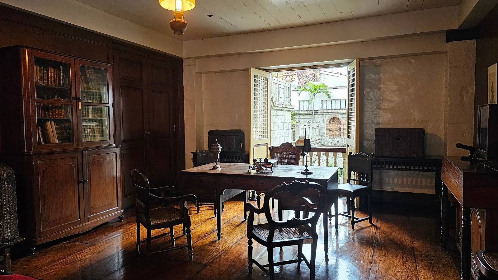 A vintage office room with wooden floors, featuring a large desk and several chairs. Bookshelves filled with books line the left wall. Sunlight streams in through a partially open window with shutters, creating a cozy and warm atmosphere.