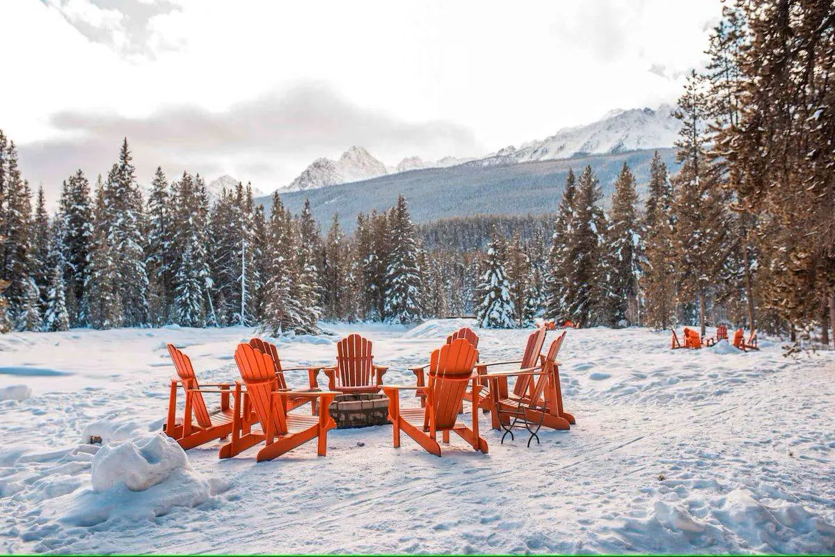 A snowy landscape with a group of orange Adirondack chairs arranged around a fire pit. Snow-covered trees surround the area, and mountains are visible in the background. Another set of chairs is in the distance.