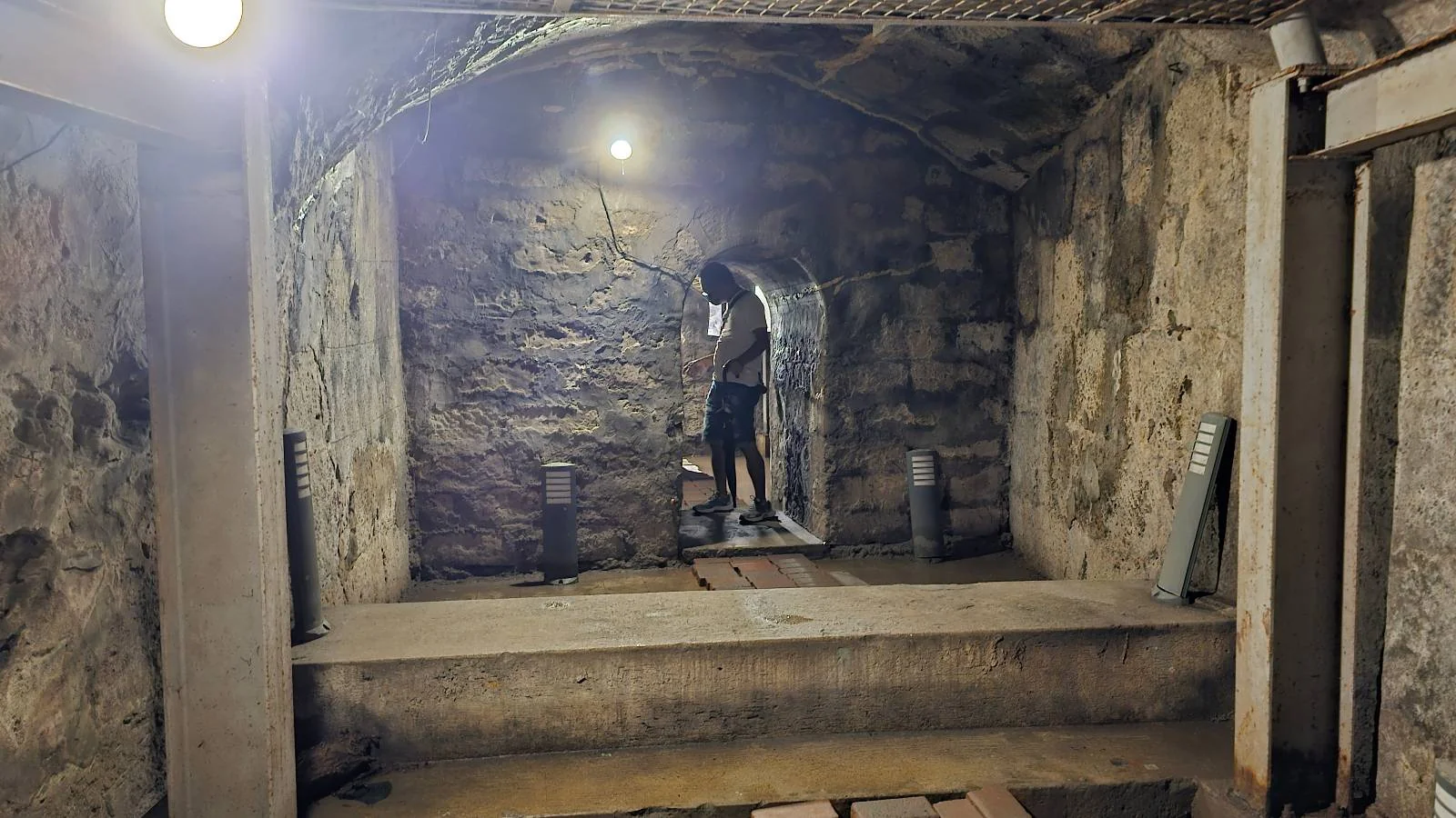 A person examines a stone wall in an underground tunnel or basement. The area is dimly lit by a single overhead light, and there are wooden planks on the sandy ground. The walls and ceiling are made of roughly hewn stone.