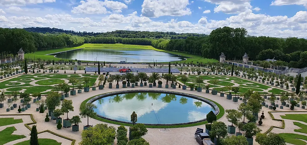 Formal gardens with ornate hedges, a circular pond, and rows of potted plants. In the background, a lake beneath a cloudy sky.