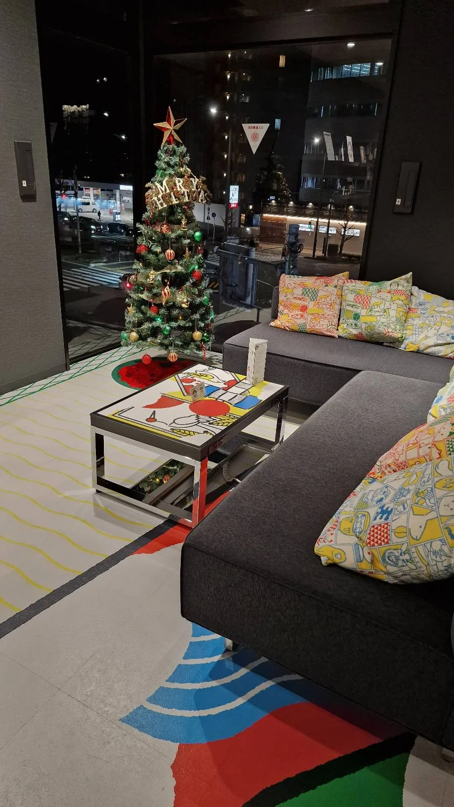 A cozy room with a modern black couch, colorful cushions, and a small Christmas tree decorated with lights and ornaments. The coffee table holds books and decor. Large windows show a view of city lights at night.