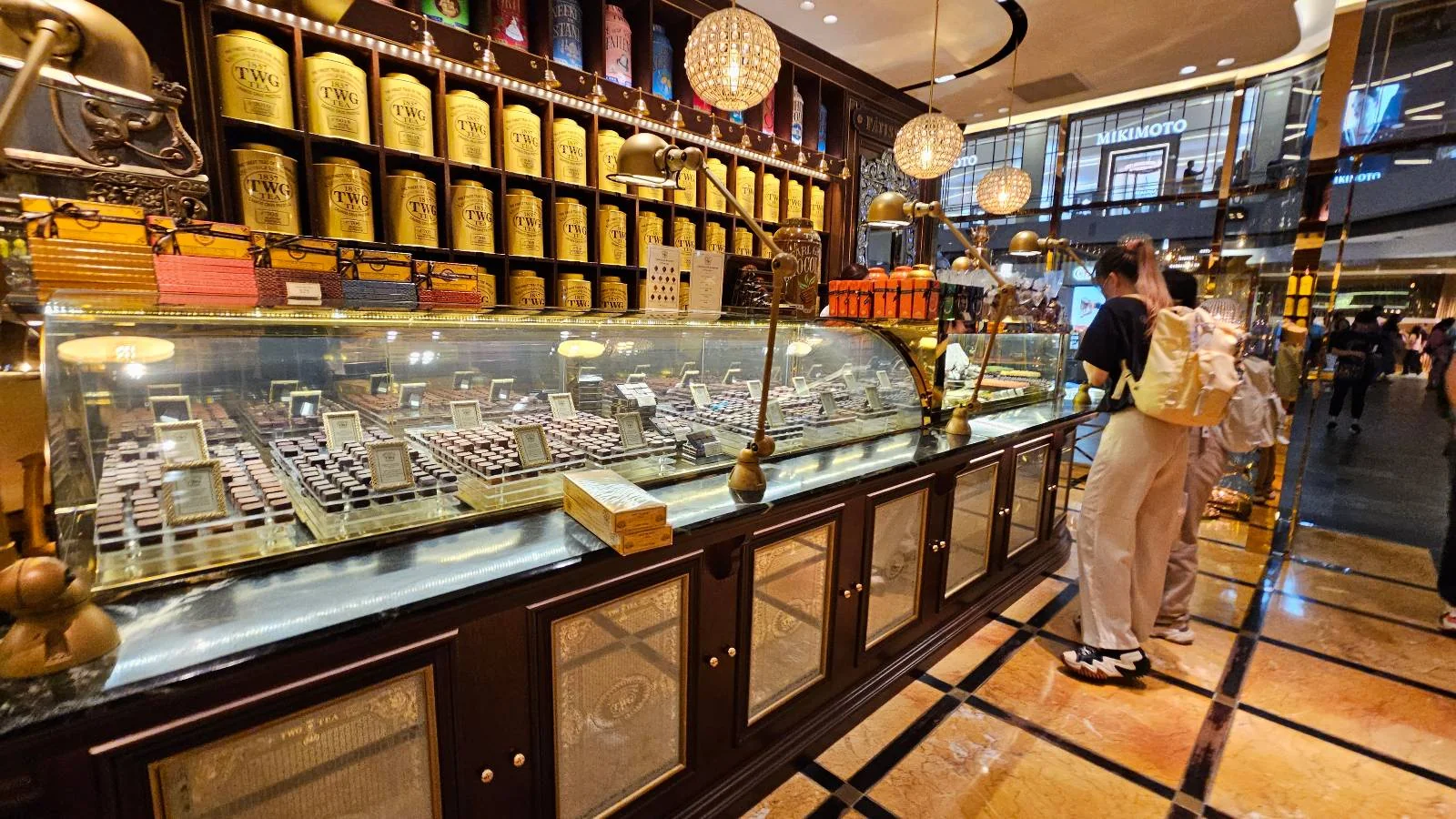A person stands in front of a pastry display inside a bakery. The counter is lined with various pastries under glass, and the wall behind features stacks of gold-colored tins. The floor features a geometric pattern, adding to the elegant interior design.