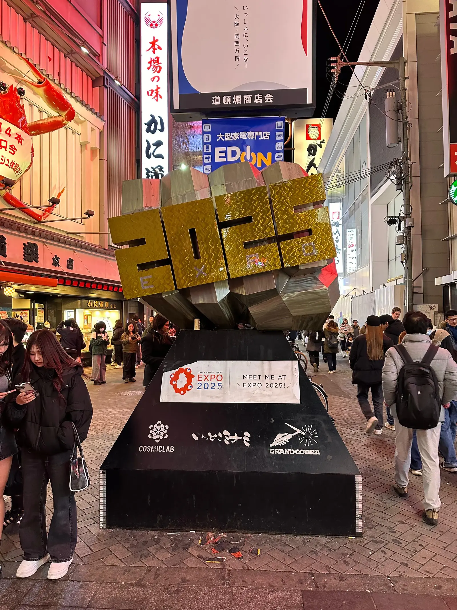 A golden "2025" sculpture is displayed on a black platform along a busy, brightly lit urban street with people walking and signs in Japanese visible in the background.
