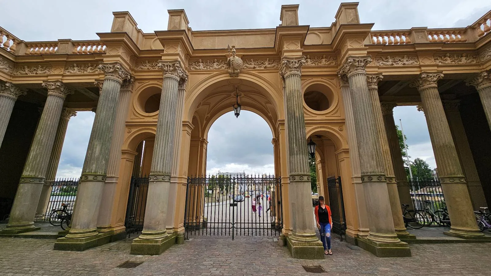 A grand stone archway with tall columns and ornate details, featuring a central iron gate. People walk nearby, and a cloudy sky looms overhead.