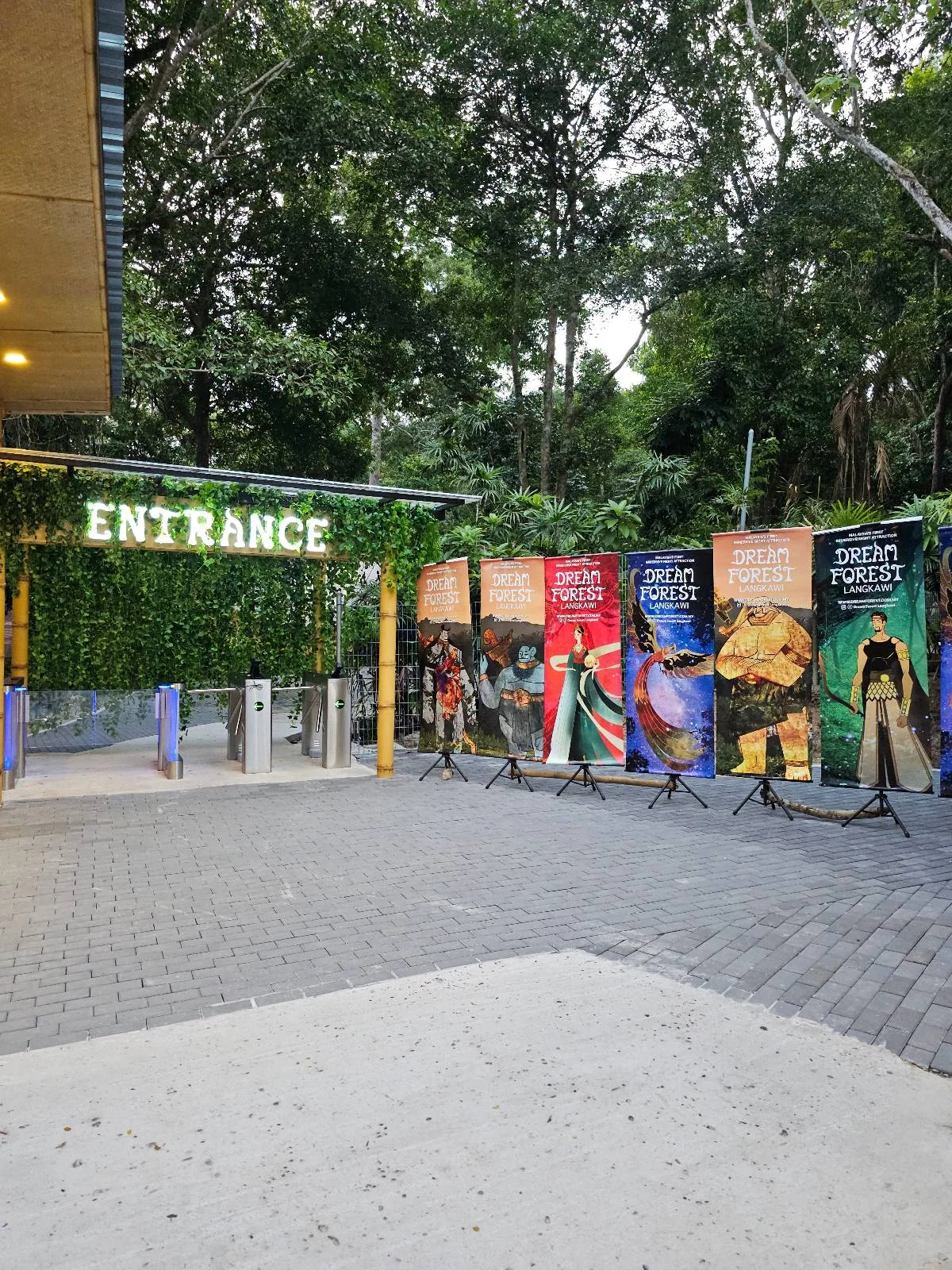 Entrance to a park with a lit "Entrance" sign. Colorful banners showcasing various foods are displayed nearby. The area is surrounded by dense green trees, and a pavement leads into the park.