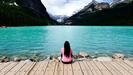 A person sitting on a wooden dock gazes at the crystal-clear turquoise waters of Lake Louise, surrounded by the majestic Rocky Mountains under a partly cloudy sky.