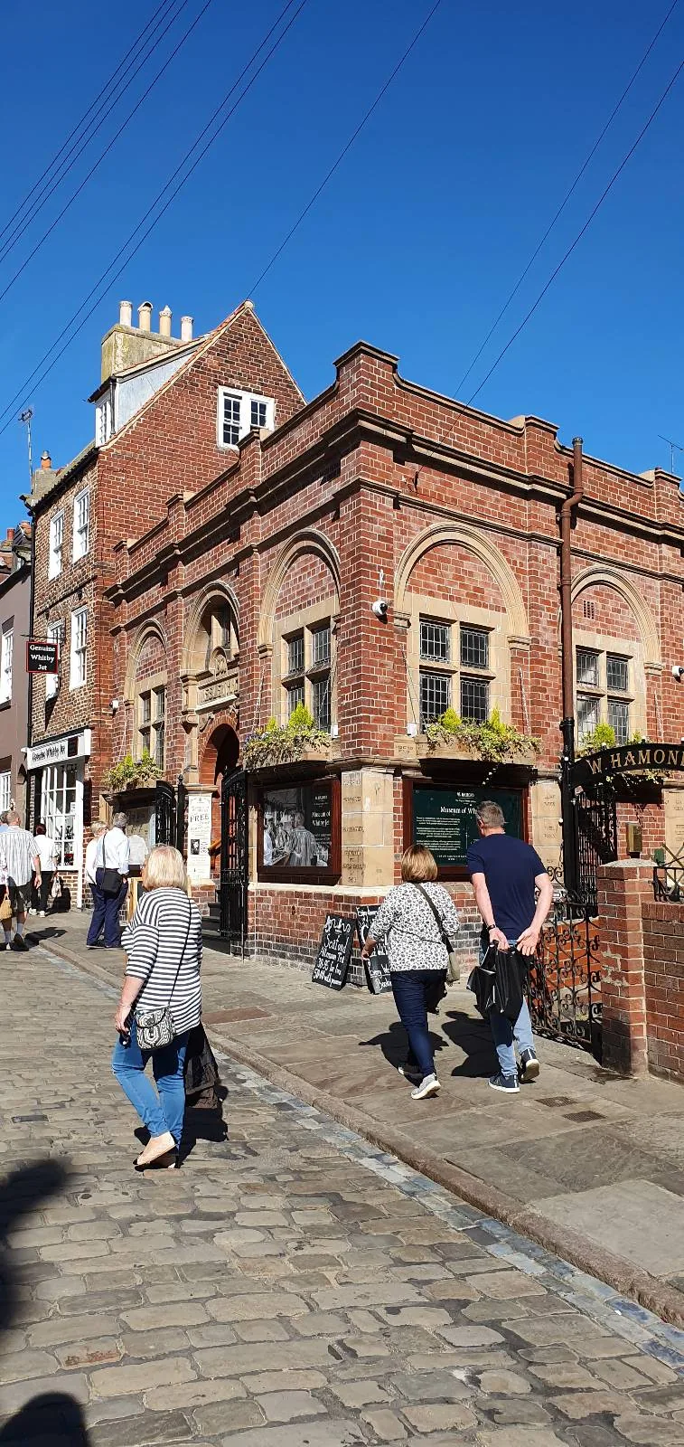 A red-brick building with ornate windows and hanging flower baskets under a clear blue sky. People are walking on the cobblestone street in front. The architecture suggests a historical or traditional pub.