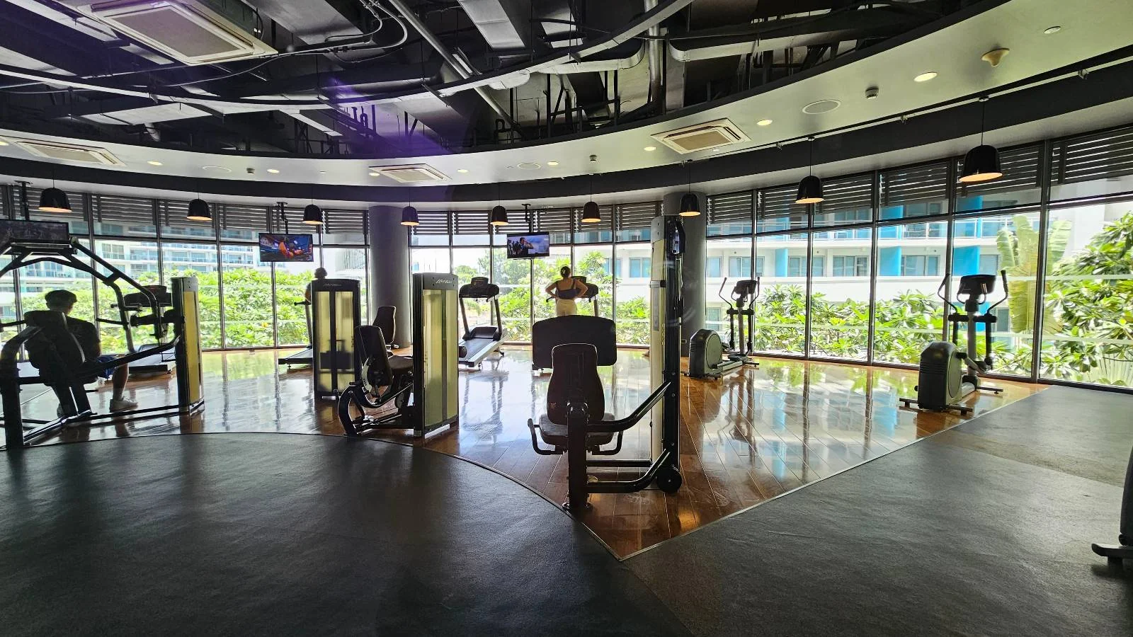 A modern gym with various exercise machines, large windows letting in natural light, and a view of greenery and buildings outside. The gym has a sleek, spacious design with polished floors and overhead lighting.
