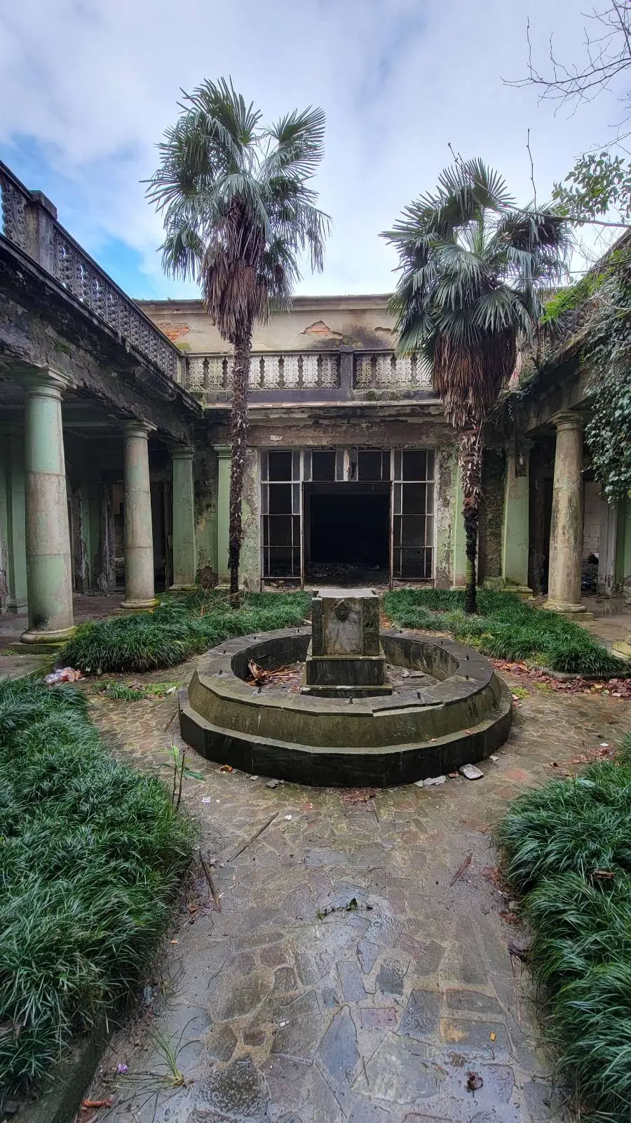 Dilapidated courtyard with overgrown plants and a central fountain, flanked by decaying columns and an abandoned building.