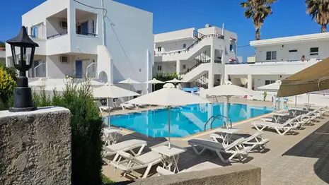Outdoor swimming pool at Michalis Apartments in Greece, with white buildings, sun loungers, umbrellas, and palm trees under a clear blue sky.