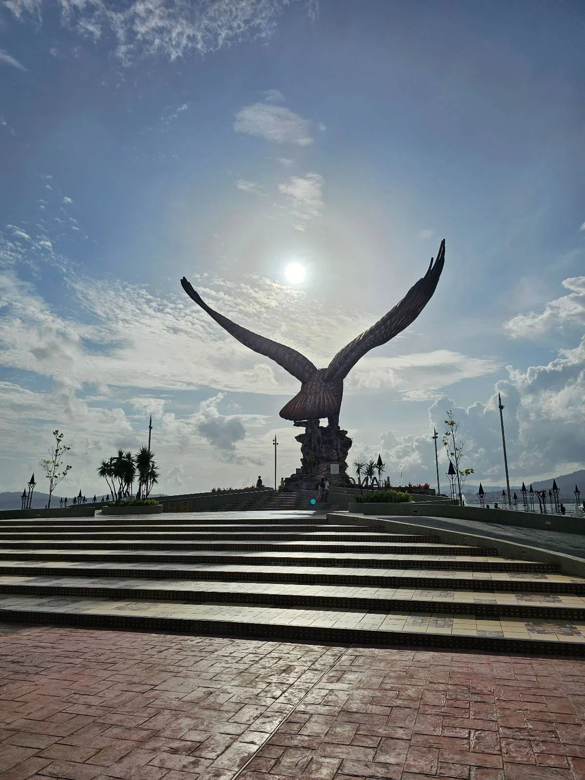 A large eagle statue with wings spread stands on a pedestal under a bright sun, surrounded by steps and a paved area, with a clear blue sky and scattered clouds in the background.