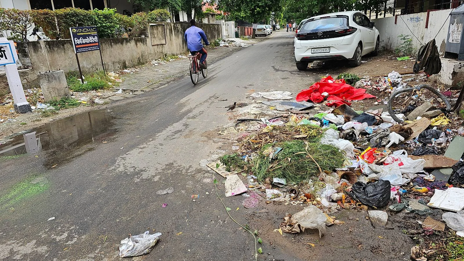 A street with scattered garbage and litter on the roadside, a white car parked nearby, and a person riding a bicycle in the background. The area appears neglected, with puddles and overgrown vegetation visible.