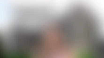 Woman smiling in front of a stone ruin with arched windows, wearing sunglasses, arms outstretched. Text: "The Ruins That Raised Me."