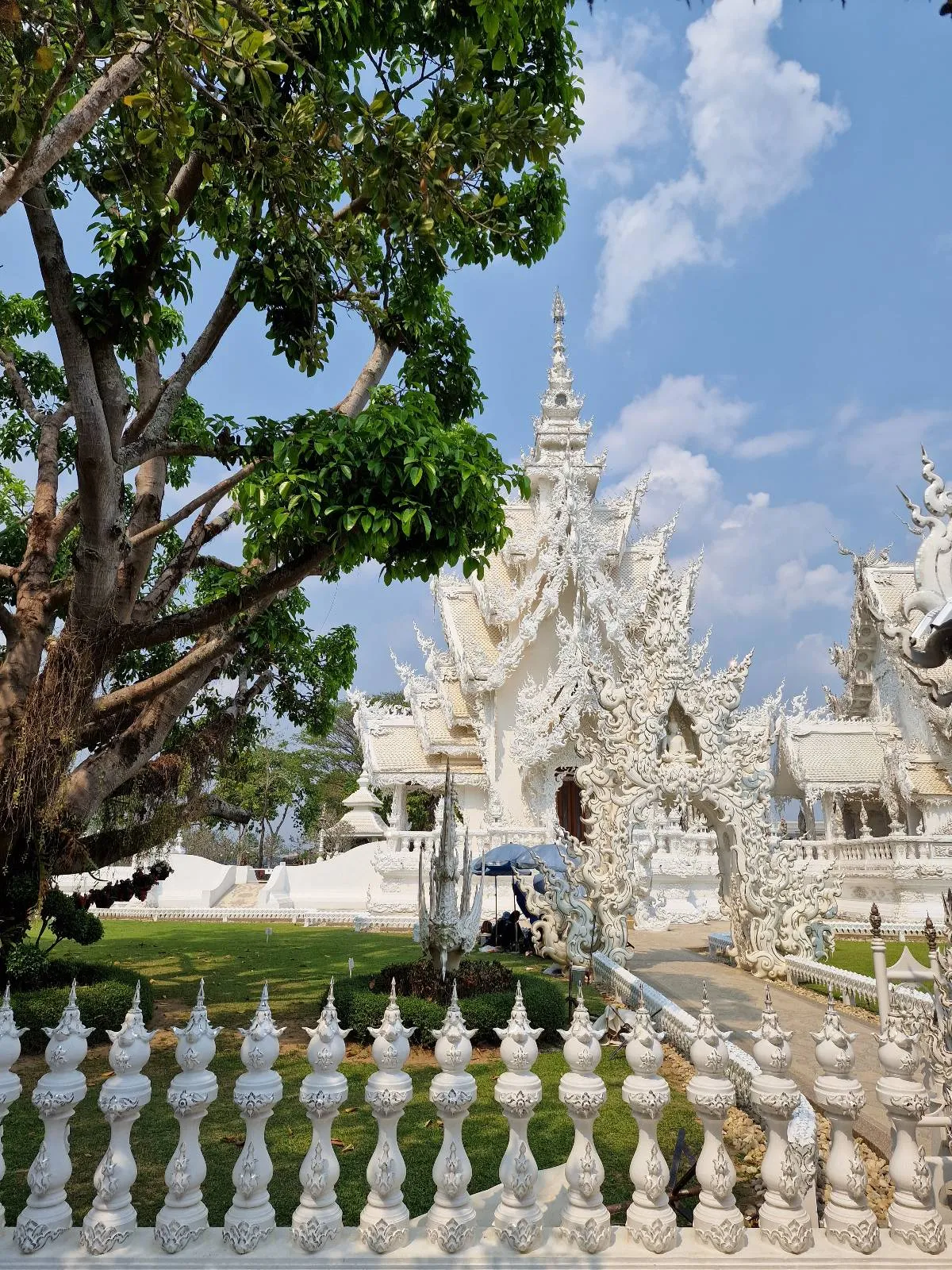 A stunning white temple with intricate carvings and ornate spires, set against a blue sky. Lush green trees frame the scene, and a decorative white fence is in the foreground. The architecture is detailed and elegant.