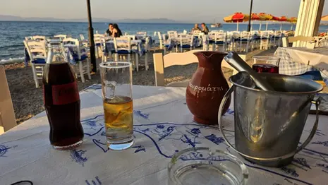 Table at Mavromatis Taverna in Greece with drinks, a water jug, and ice bucket, set beside the sea with beachfront dining tables and umbrellas in the background.