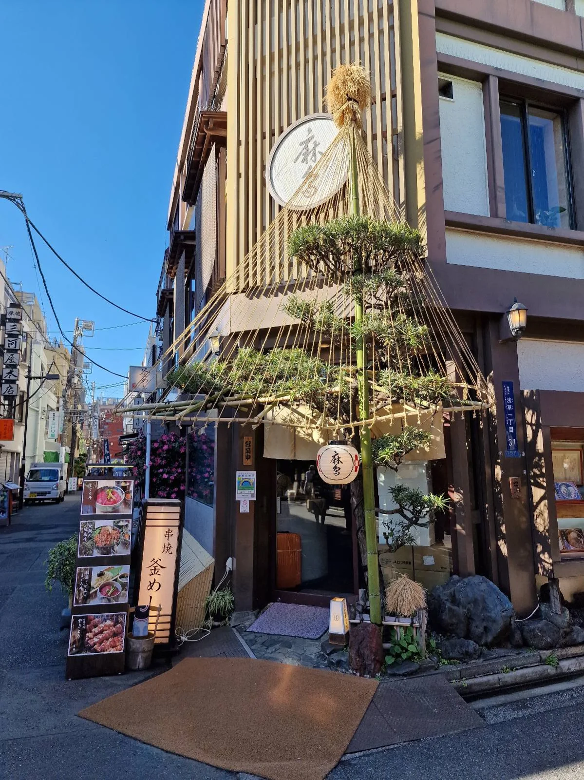 Street view of a Japanese restaurant with a wooden facade and a decorated entrance. A pine tree with protective straw wrapping is in front, alongside a menu display. A blue sky and neighboring buildings are visible in the background.