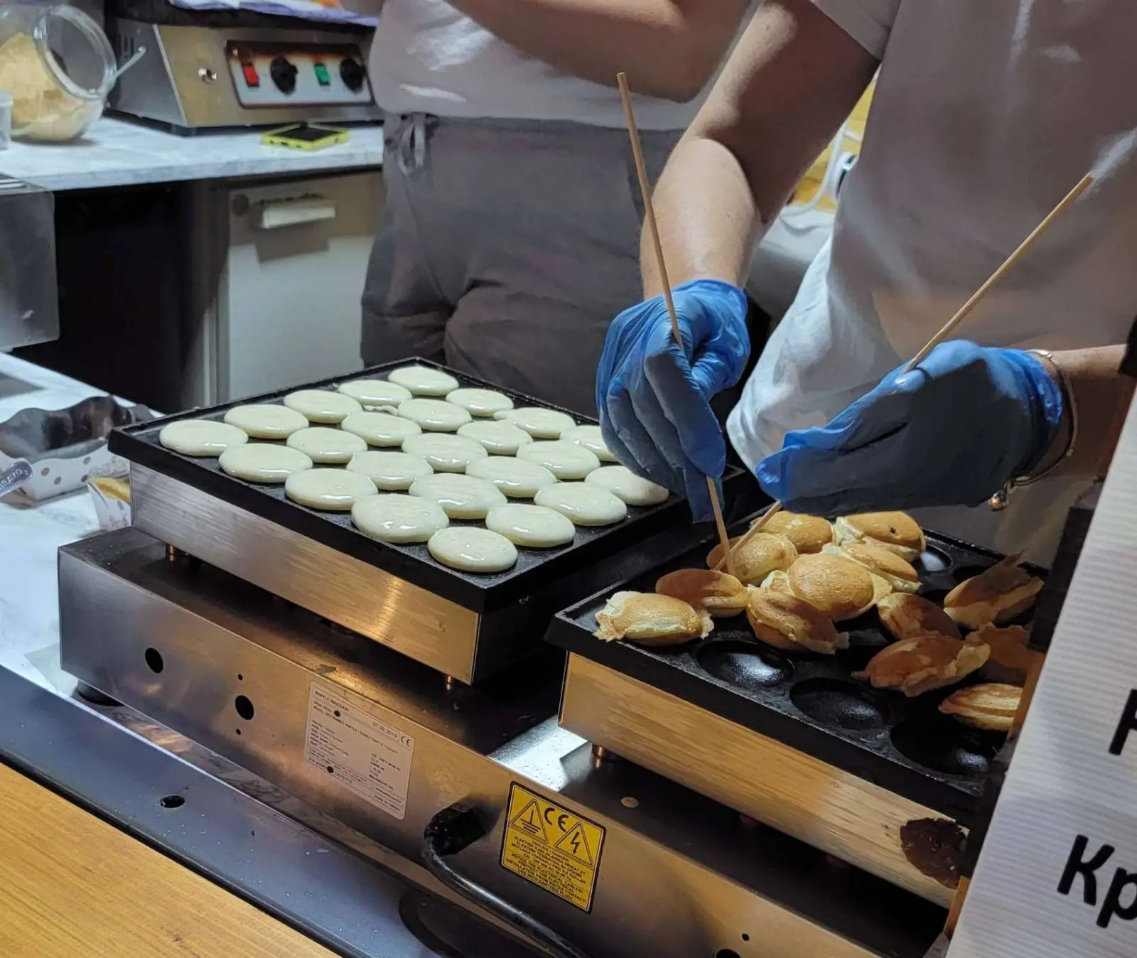 A person wearing blue gloves is using wooden skewers to flip small circular pastries on a griddle. A tray of uncooked pastries is on the left, while cooked ones are on the right. Kitchen equipment is visible in the background.