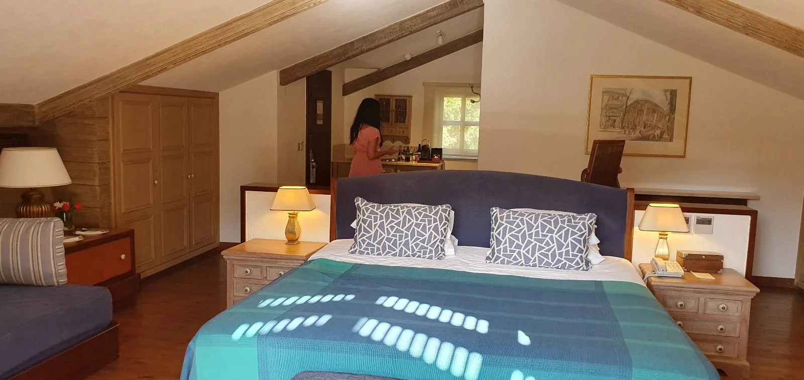 A cozy attic bedroom with exposed wooden beams. The room features a double bed with blue bedding and patterned pillows, wood nightstands with lamps, a sofa, and a window in the background. A person stands near a dresser.