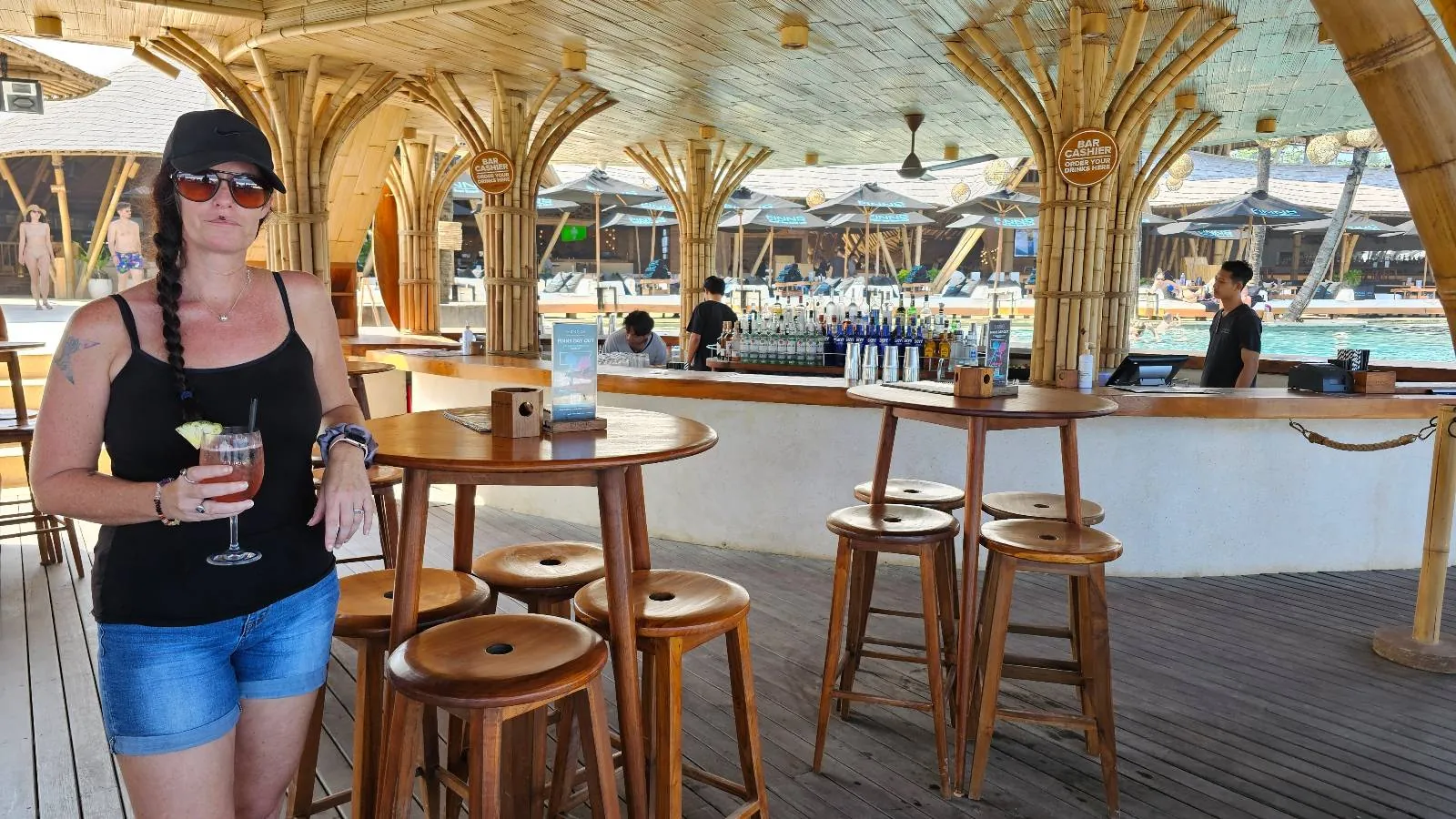A woman in a black tank top and sunglasses stands at a tropical beach bar holding a cocktail. The bar is made of bamboo and has tall stools. In the background, there's a pool and umbrellas under a sunny blue sky.