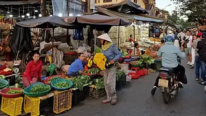 A busy street market scene in Hoi An with vendors selling fresh green vegetables and a person in a conical hat carrying bright yellow flowers.