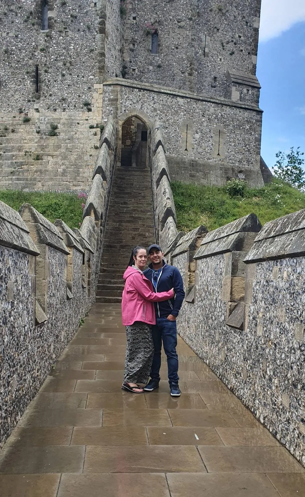 A couple embraces while standing on a stone walkway with high walls on either side, leading to an old stone building. Steps ascend to a doorway behind them. The sky is clear with a few clouds.