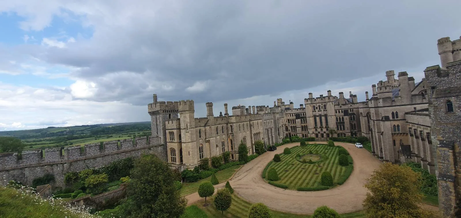 A panoramic view of a sprawling medieval castle with towers and battlements set amidst greenery. The castle's courtyard features a circular garden path. The sky is cloudy, creating a dramatic backdrop.