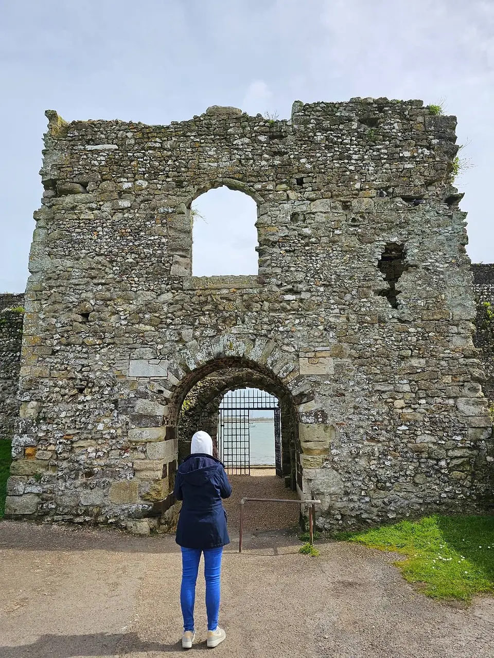 A person in a blue jacket and white headscarf stands facing an ancient stone archway with a view of the sea through it.