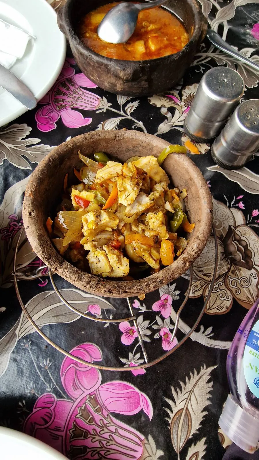 A bowl filled with a colorful vegetable and meat dish sits on a floral-patterned tablecloth. Nearby are a metal spoon, a silver salt and pepper shaker, and a wooden bowl with a bright orange sauce.