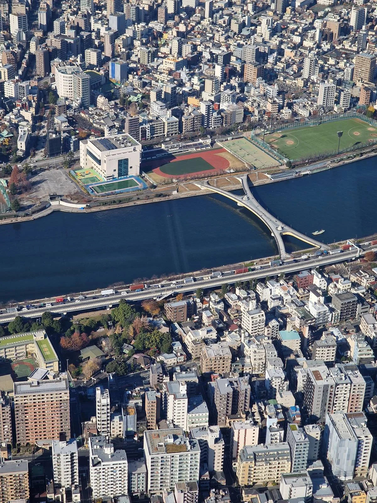 Aerial view of a sprawling cityscape with densely packed buildings. Two rivers run parallel through the city, with multiple bridges connecting the areas. In the distance, a mountain peak is visible under a clear blue sky.