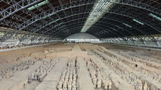 Rows of life-sized Terracotta Warriors stand in an excavation pit under a large arched metal roof, with visitors observing from walkways along the sides of the structure.