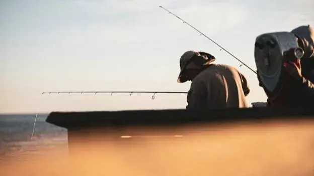 Two people fishing at sunset by the sea, beige and blue tones, one handles a rod, the other examines gear, creating a calm atmosphere.