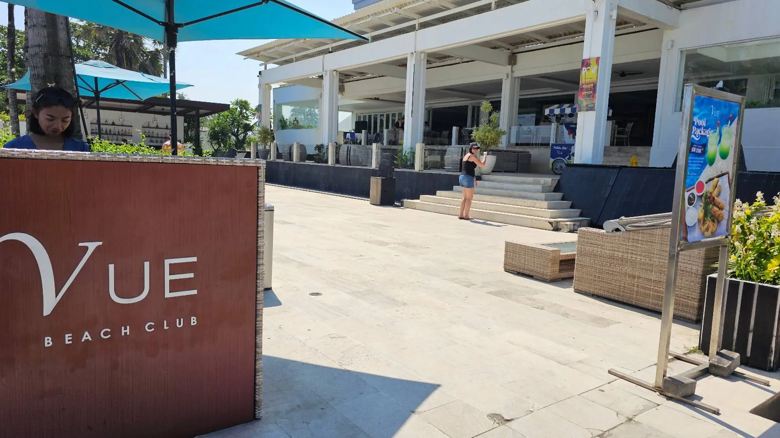 Vue Beach Club entrance with a woman standing beside a counter and another person taking photos. Blue umbrellas, pool package sign, sunny day.