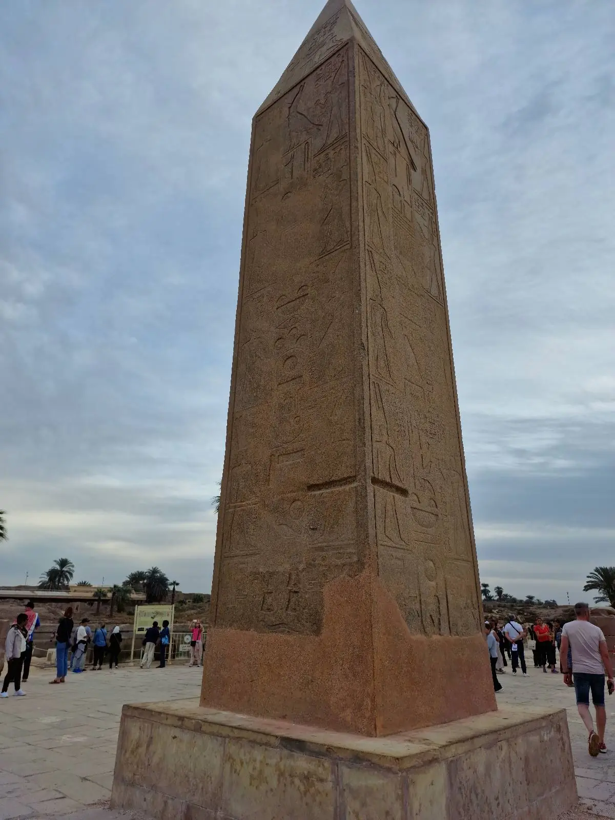 A tall, ancient stone obelisk stands under a cloudy sky, surrounded by tourists. The obelisk is adorned with carvings and hieroglyphics.
