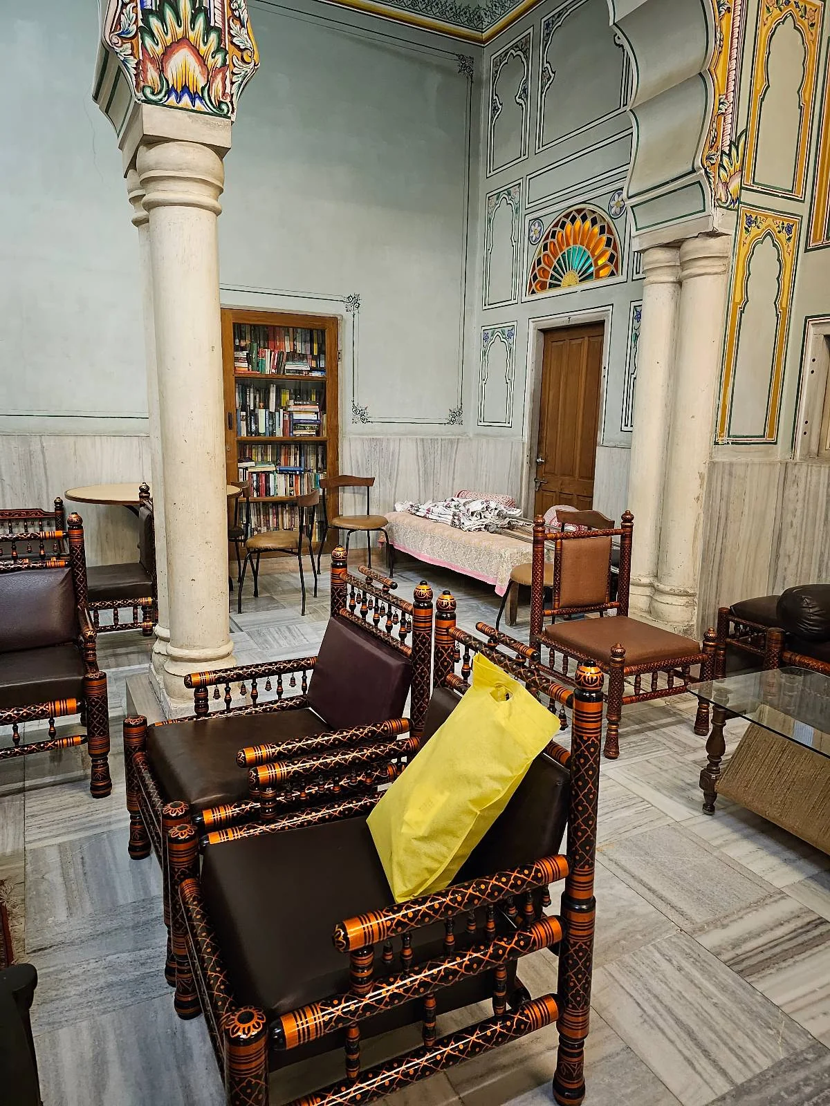 A room with ornate columns, wooden chairs with cushions, a table, bookshelves, and colorful arch decorations on the walls. The floor is tiled with light-colored marble.