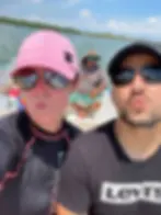 Two people in sunglasses make kissy faces on a boat, wearing a pink hat and a Levi's shirt. A man sits behind them. Sunny day, calm water.