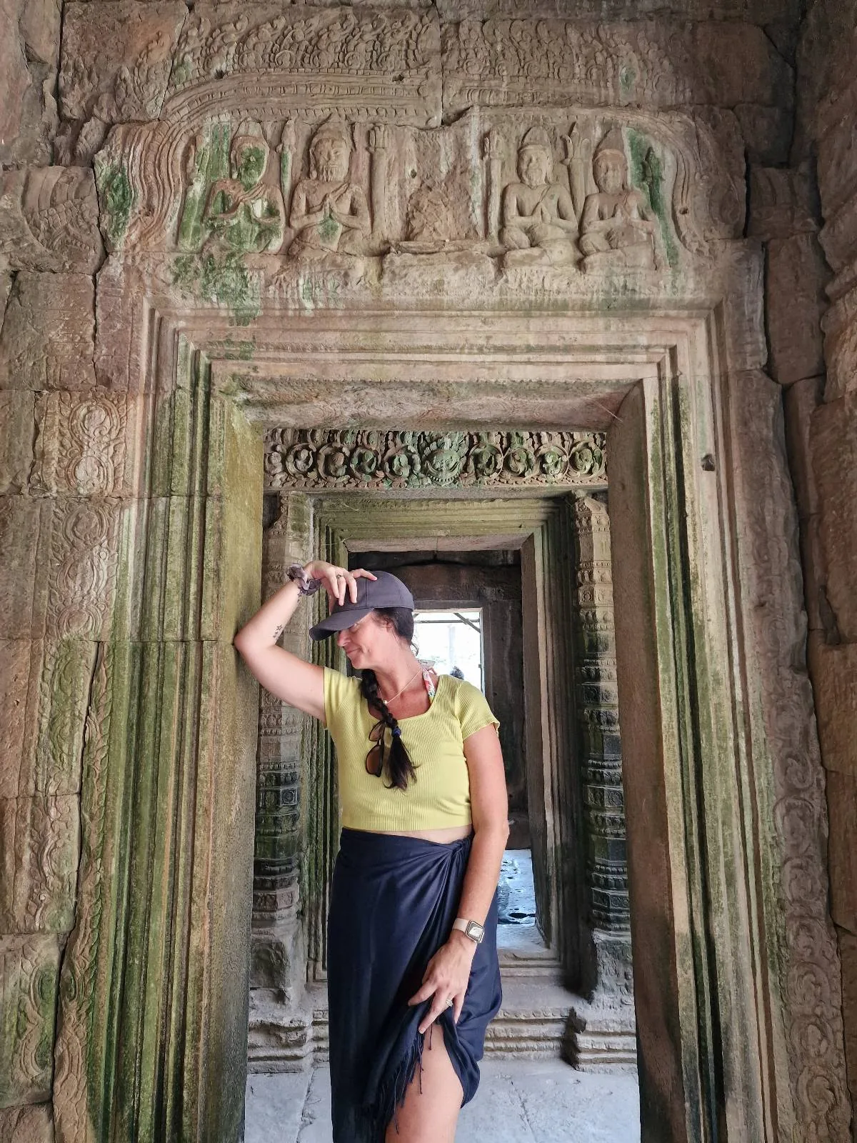 A woman stands posing under an ancient stone archway with intricate carvings, surrounded by weathered walls and moss, in what appears to be a historical temple or ruin.