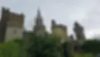 View of a medieval castle with multiple towers and spires, surrounded by lush greenery under a cloudy sky.
