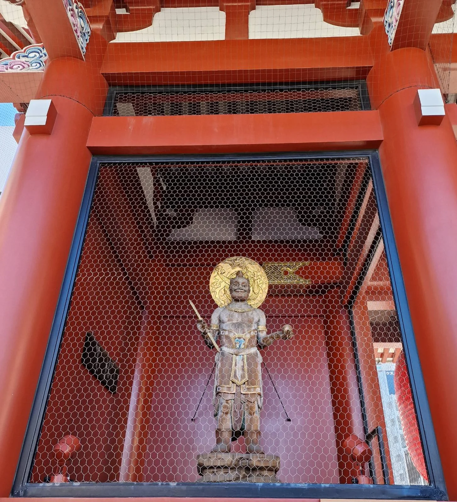 Stone statue with a golden halo behind it inside a red shrine with a mesh barrier. Intricate patterns adorn the pillars. Bright, serene setting.