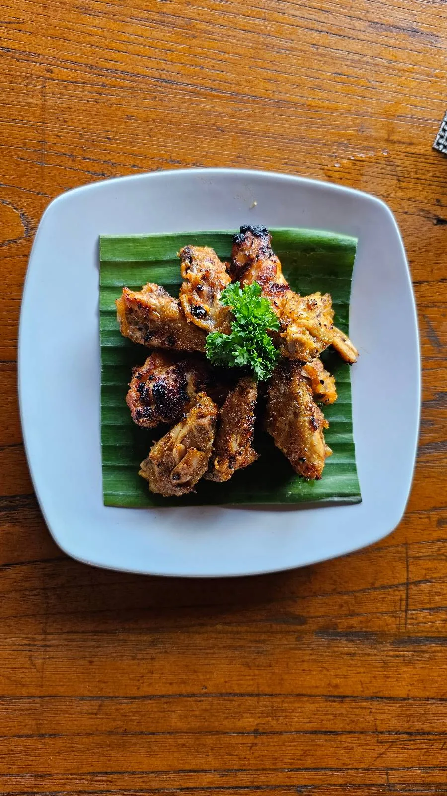 Outdoor restaurant with wooden tables and chairs, surrounded by tropical plants. The seating area is covered with a wooden roof, creating a cozy and inviting atmosphere. The decor includes colorful artwork and natural elements.