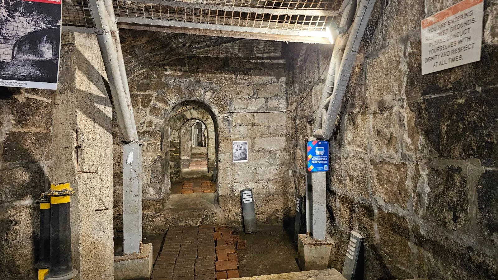 A narrow, dimly lit stone corridor with multiple archways. Metal supports line the walls, and various signs are posted. The floor is wooden, leading deeper into the passage.