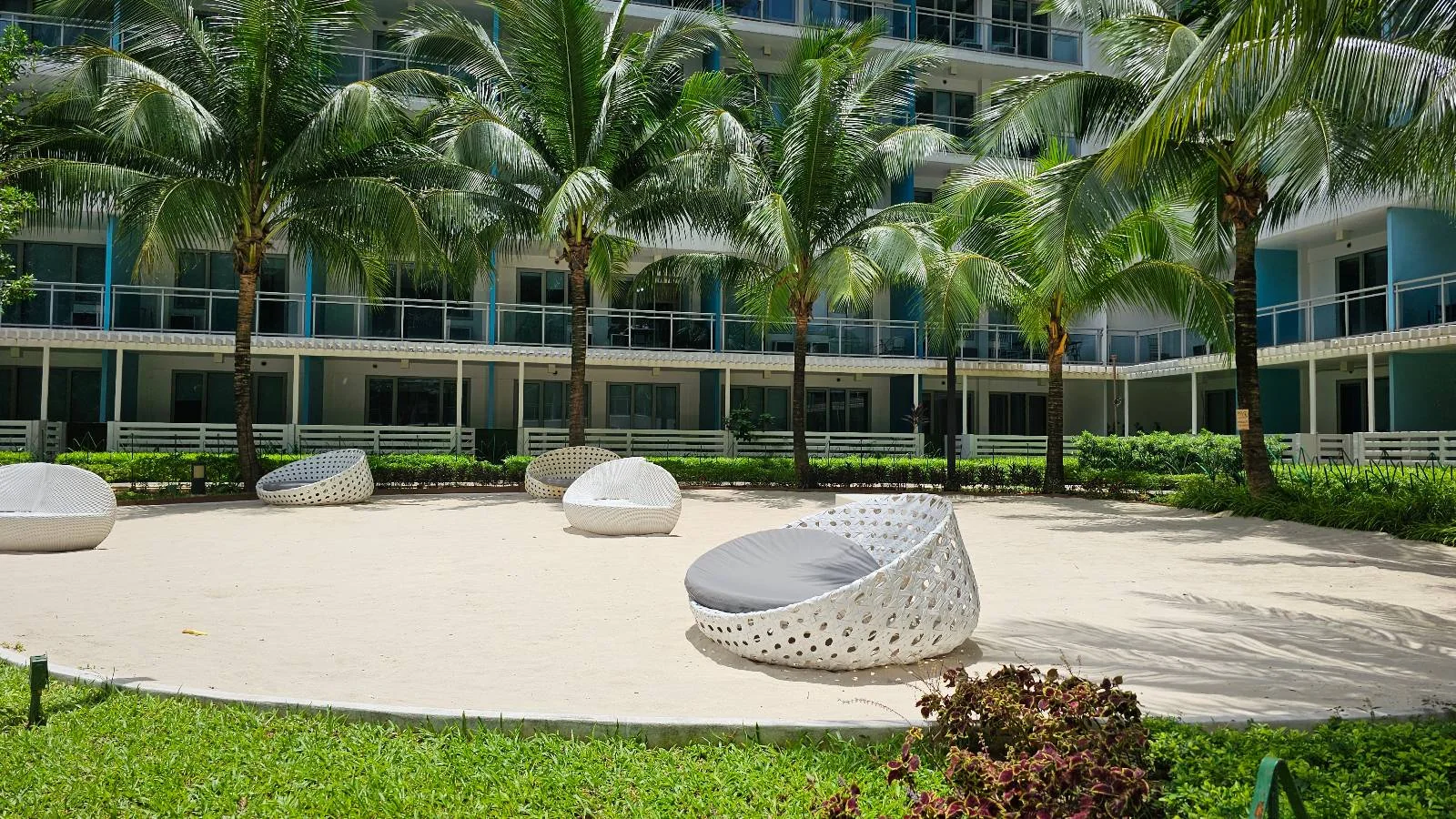 White outdoor lounge chairs are arranged on a sandy area surrounded by green grass, palm trees, and a modern multi-story building with balconies in the background.