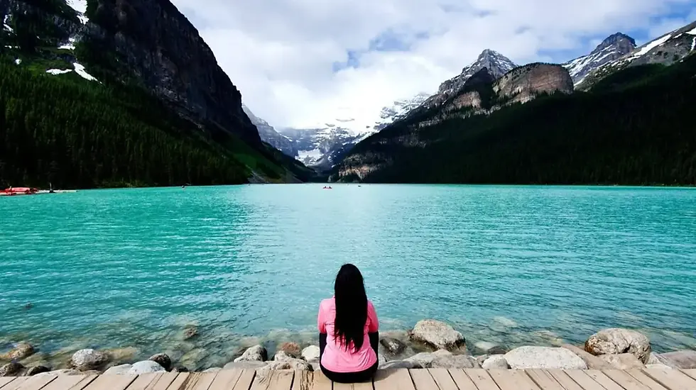 Woman in a pink top sits on a dock by a turquoise mountain lake, facing snowy peaks and a calm, scenic valley.