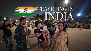 Crowd in a well-lit night street in India. A woman holds a baby, smiling adults converse. Text reads "Traveling in India" with a flag motif.