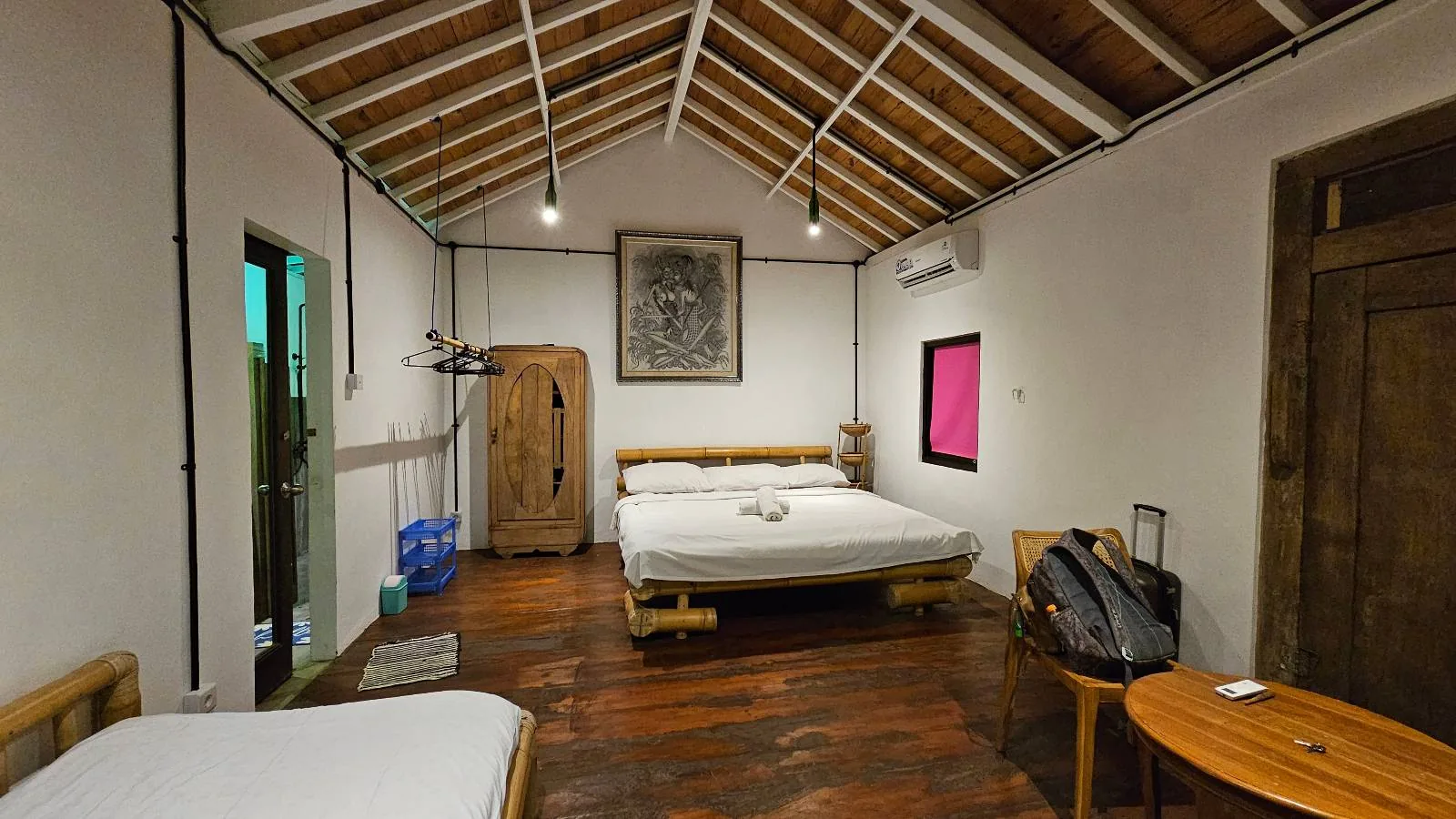 Cozy bedroom with a slanted wooden ceiling, double bed, and a painting. A backpack rests on a chair, and a pink curtained window.