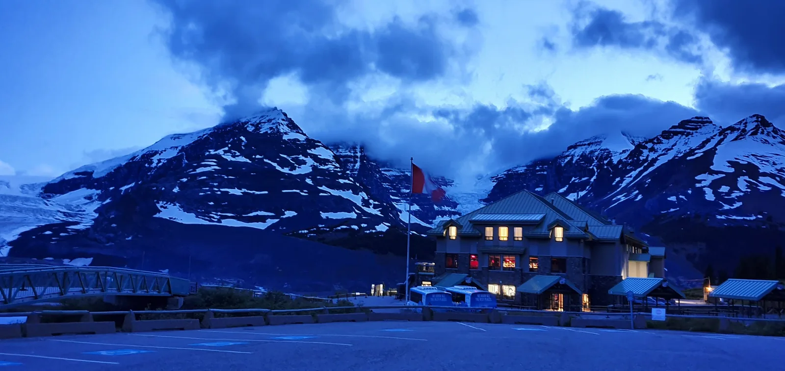 A building with lit windows stands in the foreground under a cloudy, twilight sky. Snow-capped mountains loom in the background, creating a dramatic contrast. Some flags are visible near the building, and the scene has a tranquil, cool ambiance.