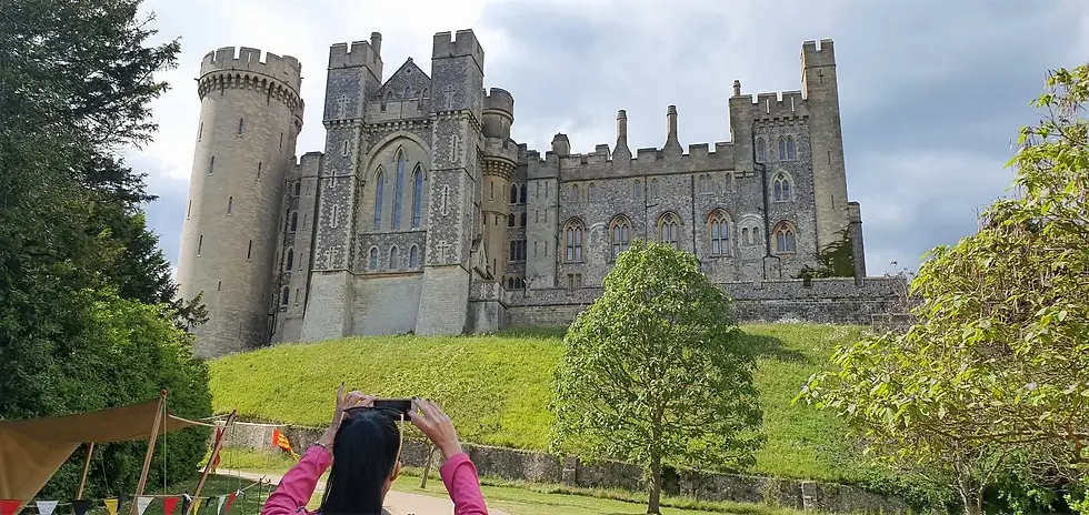 Person in pink taking a photo of a large stone castle with towers. Greenery and trees surround the setting under a cloudy sky.